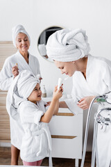 Smiling kid in bathrobe holding cream near lesbian parent in bathroom