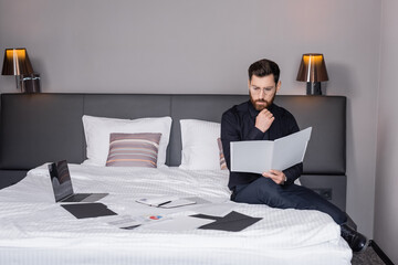 pensive man in suit and eyeglasses looking at folder and sitting near laptop on hotel bed