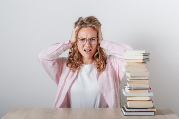 A girl in stress holds her head and looks at a large stack of books
