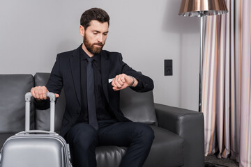 bearded businessman checking time on wristwatch and sitting near luggage in hotel room