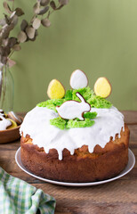 Easter cake with raisins decorated with white icing and gingerbread in the shape of Easter eggs and a rabbit on green grass on a wooden table. Festive pastries for a family dinner. Rustic.