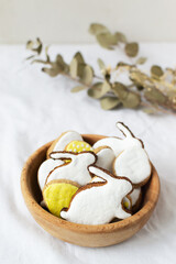 Festive pschal gingerbread in the form of eggs and rabbits with colored icing in a bowl on a white background with willow branches. The concept of traditional holiday food. Vertical orientation.