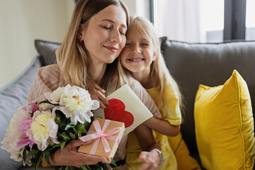 Child daughter congratulates mother and gives present card, gift and bouquet of flowers at home. Mom and girl smiling and hugging on couch. Happy mothers day celebration