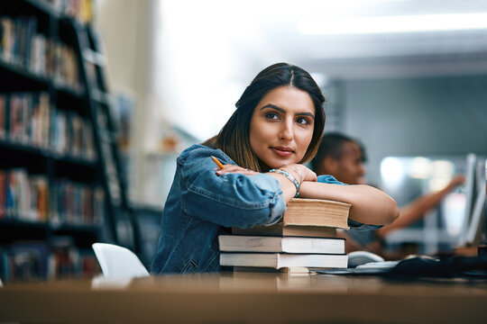 Where dreams are turned into reality. Shot of a young woman resting on a pile of books in a college library and looking thoughtful.
