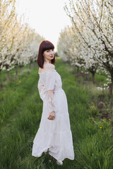 Full length portrait of gorgeous dark haired woman wearing white long dress standing a spring garden with blooming apple trees. Leisure activity on fresh air of caucasian young lady.