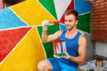 The guy paints the wall with a brush in bright colors, holds a can of paint in his hand