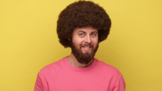 Positive bearded man with Afro hairstyle blinking eye joyfully as if having interesting idea, looking playfully flirting, wearing pink sweatshirt. Indoor studio shot isolated on yellow background.