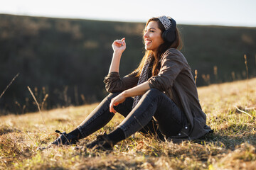 Naklejka na ściany i meble Cute smiling young woman listening music on her headphones while sitting on the ground in early spring sunny day and enjoying calm.