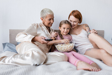 Smiling girl holding popcorn while watching tv near lesbian moms at home