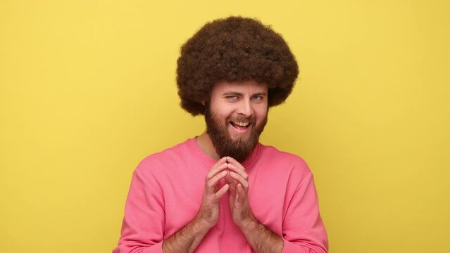 Bearded man with Afro hairstyle thinking devious plan with cunning face expression, having slyly idea to prank, wearing pink sweatshirt. Indoor studio shot isolated on yellow background.