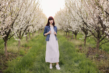 Obraz premium Full length portrait of charming young woman wearing white dress and denim jacket spending leisure time among beautiful blooming garden. Concept of people, beauty and relaxation.