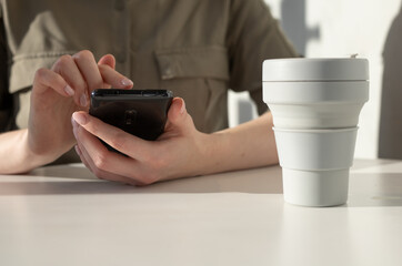 Female hands closeup holding phone. Woman sitting at table with collapsible cup and using smartphone for relax, work or during coffee break. High quality photo
