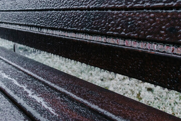 A wooden brown bench covers a layer of ice and icicles after freezing rain in winter