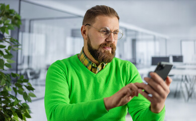 Fototapeta premium Focused bearded man in bright green sweater and eyeglasses using modern smartphone while working at bright office. Concept of people, business and gadgets.