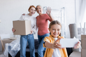 Positive kid having video call on smartphone near mothers with carton box at home