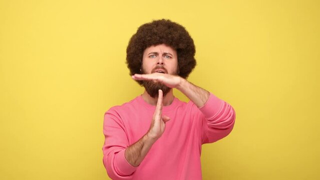 I need more time. Bearded man with Afro hairstyle showing time out gesture, looking at camera, hurry to meet deadline, wearing pink sweatshirt. Indoor studio shot isolated on yellow background.