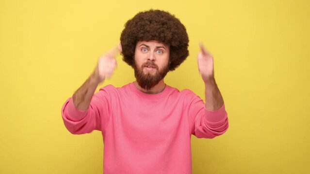 Must be here and right now. Bearded man with Afro hairstyle standing, pointing down and looking at camera, wearing pink sweatshirt. Indoor studio shot isolated on yellow background.