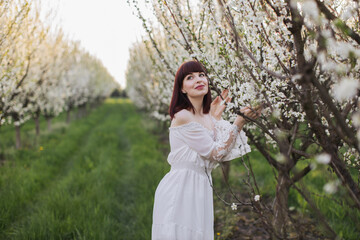 Fototapeta na wymiar Adorable caucasian woman with dreamy looking standing near apple tree and touching gently fresh blossom. Young lady with brown hair enjoying free time among spring nature.
