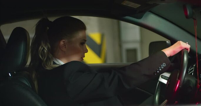 a beautiful blonde woman in a black suit, white shirt and red tie sits in her black car in an underground parking lot, looking sternly to the side. cinematic frame, slow motion.