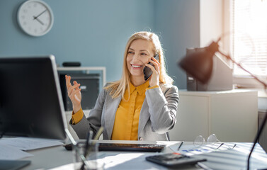 Positive mature woman in stylish formal wear talking on mobile phone while sitting at office desk...