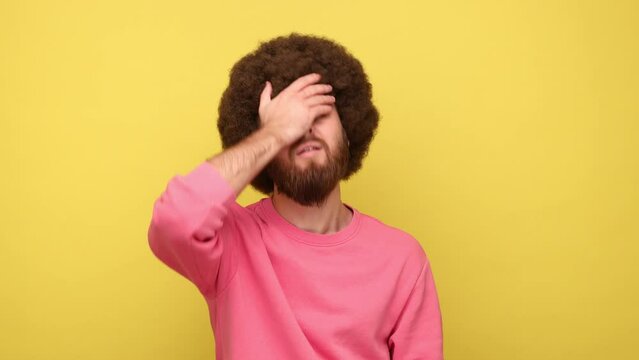 Bearded man with Afro hairstyle standing with facepalm gesture, blaming himself, feeling sorrow regret because of bad memory, wearing pink sweatshirt. Indoor studio shot isolated on yellow background.