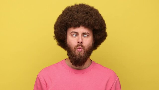 Bearded man with Afro hairstyle looking around with crossed eyes, making silly brainless face, looks stupid, aping as fool, wearing pink sweatshirt. Indoor studio shot isolated on yellow background.