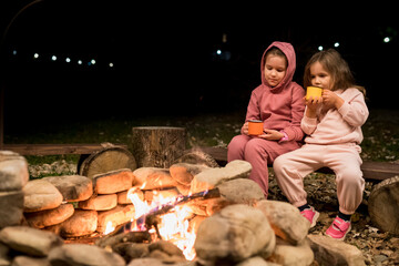 Fun camping activities for kids. Girls sitting by crackling campfire and drinking hot chocolate in the evening. Family getaway and hiking in woodland camp with children