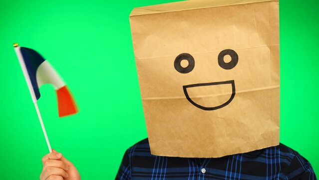 Portrait of man with paper bag on head waving French flag with smiling face against green background.