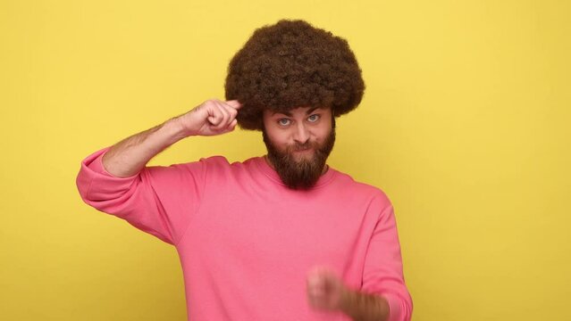 You stupid. Man with Afro hairstyle looking at camera, crossing eyes, holding finger near temple, showing stupid gesture, wearing pink sweatshirt. Indoor studio shot isolated on yellow background.