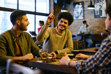 Happy Muslim man raising his hand to call a waiter while being with male friends in a pub.
