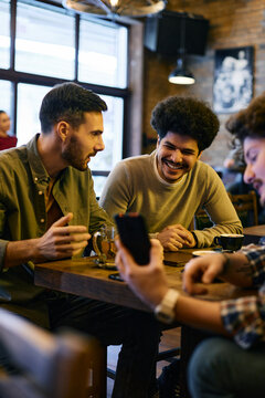 Cheerful Lebanese man and his Caucasian friends watch something on mobile phone while gathering in pub.