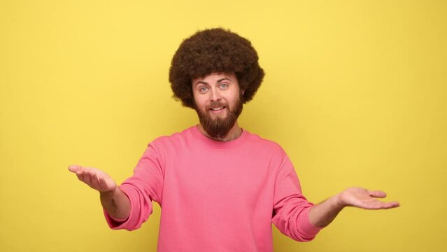 Man with Afro hairstyle sharing opening hands looking at camera with kind smile, greeting and regaling, happy glad to see you, wearing pink sweatshirt. Indoor studio shot isolated on yellow background
