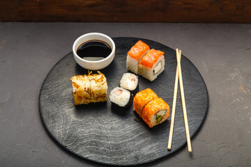 Rolls with fish and shrimp on a round board next to sticks, soy sauce on a dark background.