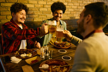 Multiracial group of male friends hav3 fun while toasting with beer and eating in pub.