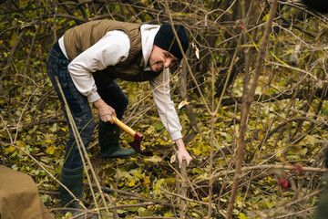 Tired tourist male wearing warm clothes chopping firewood with axe in forest on overcast cold day....