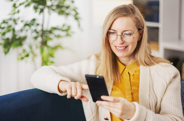 Attractive mature woman using modern smartphone while resting on cozy sofa at home. Pleasant blonde wearing comfy casual clothes and eyeglasses.