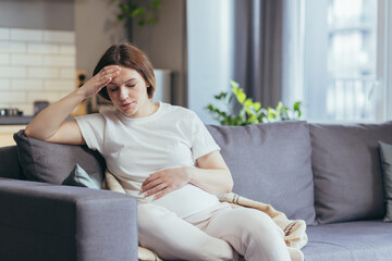 Young beautiful pregnant woman sitting at home on the couch sick, has a headache and nausea