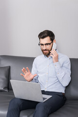smiling man in eyeglasses having phone call while sitting with laptop in hotel