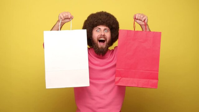 Man with Afro hairstyle raised arms with shopping bags, yelling happily, looks at camera with extremely happy expression, wearing pink sweatshirt. Indoor studio shot isolated on yellow background.