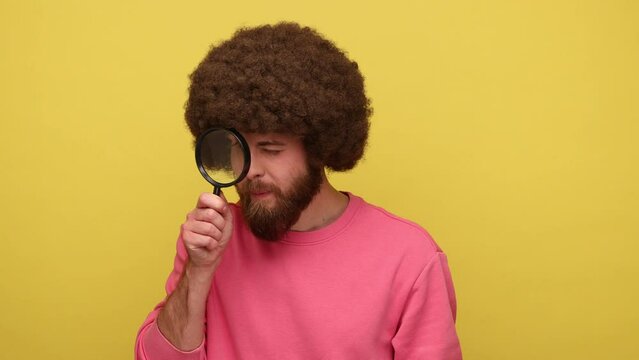 Bearded hipster man with Afro hairstyle holding magnifying glass and looking at camera with big zoom eye, wearing pink sweatshirt. Indoor studio shot isolated on yellow background.