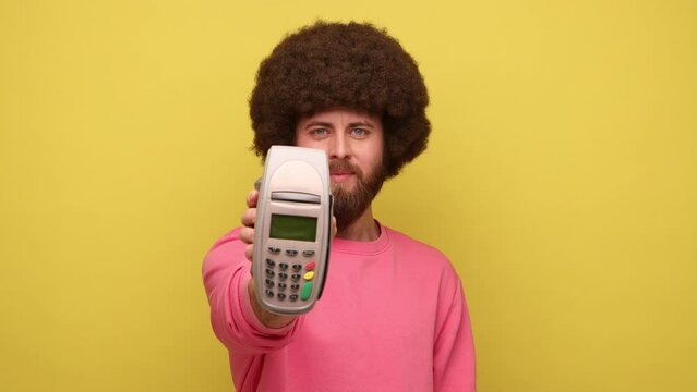 Bearded man with Afro hairstyle holding in hand pos contactless payment terminal, using paypass for purchases payment, wearing pink sweatshirt. Indoor studio shot isolated on yellow background.