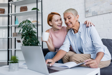 happy woman with cup of tea hugging lesbian girlfriend working on laptop at home