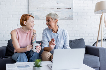 lesbian woman pointing at laptop near cheerful girlfriend with tea cup