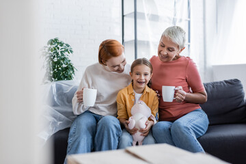 cheerful girl with toy bunny looking at camera near same sex mothers sitting with tea cups in new home