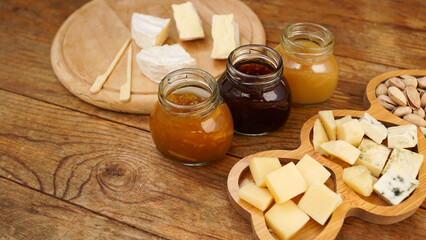 Jars of homemade jams and honey in glass jars and a variety of cheeses on a wooden table. Cheese plate for appetizers.