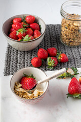 A ceramic bowl full of healthy muesli and a great strawberry piece in a spoon surrounded by much more strawberry pieces.