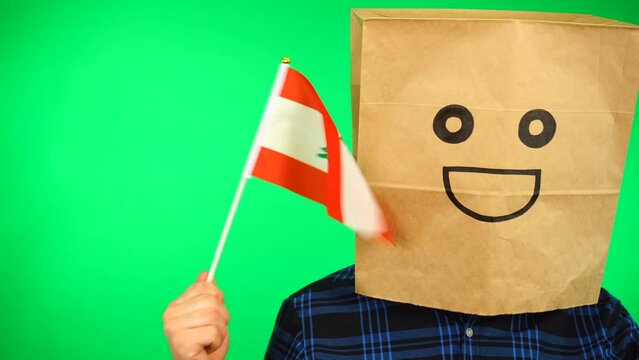 Portrait of man with paper bag on head waving Lebanese flag with smiling face against green background.