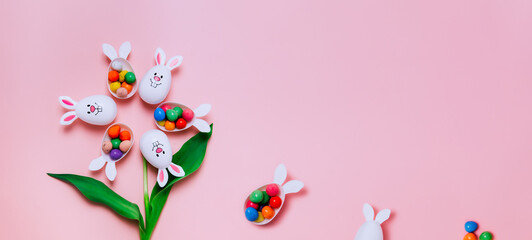 Top view Easter pink background with flower shape made of funny eggs containers with bunny ears filled with candy chocolate eggs. Bunny rabbit symbol. Easter egg hunt. Creative Easter. Wide banner.