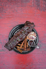 Overhead view of pork ribs marinated with barbecue sauce and herbs, sweet potato fries and coleslaw salad, in a black dish on the table.