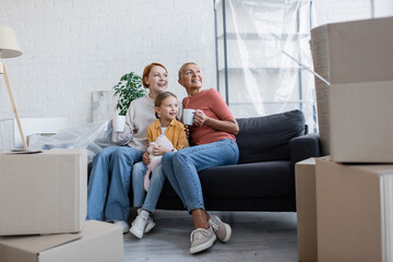 joyful lesbian women with adopted daughter looking away on couch in new apartment